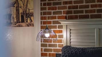 A bedroom with a brick wall, a bed with a dark cover, a hanging light fixture, and a painting on the wall.
