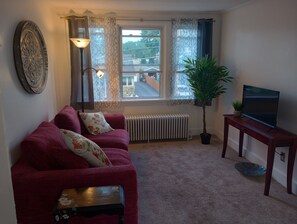 Ett vardagsrum med en röd soffa, en tv på ett träställ, ett element och ett fönster med gardiner.