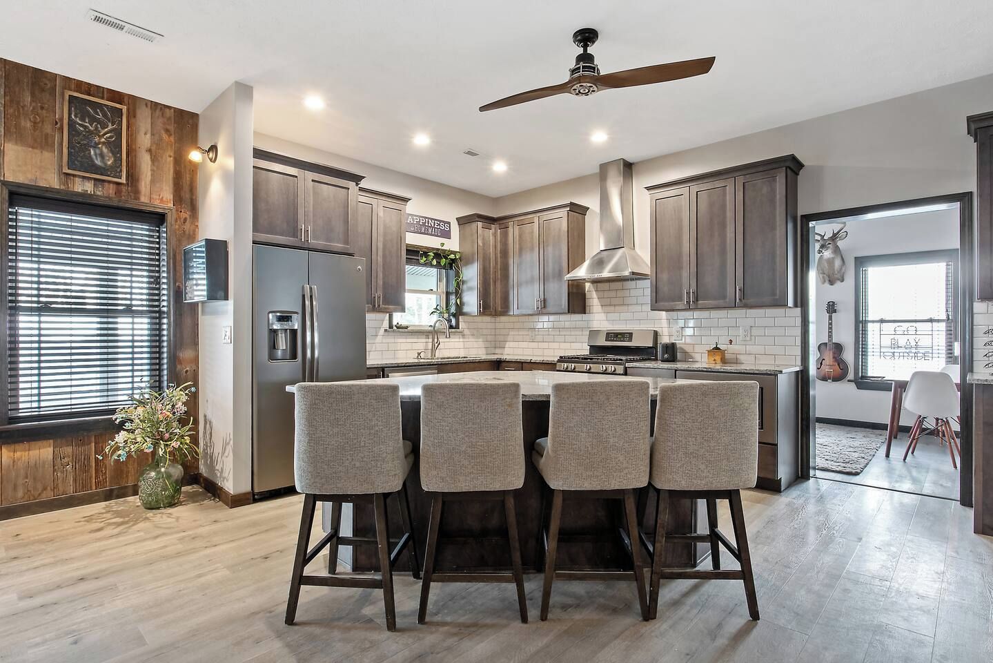A modern kitchen with a central island, bar stools, a ceiling fan, and a view of a guitar in the adjacent room.