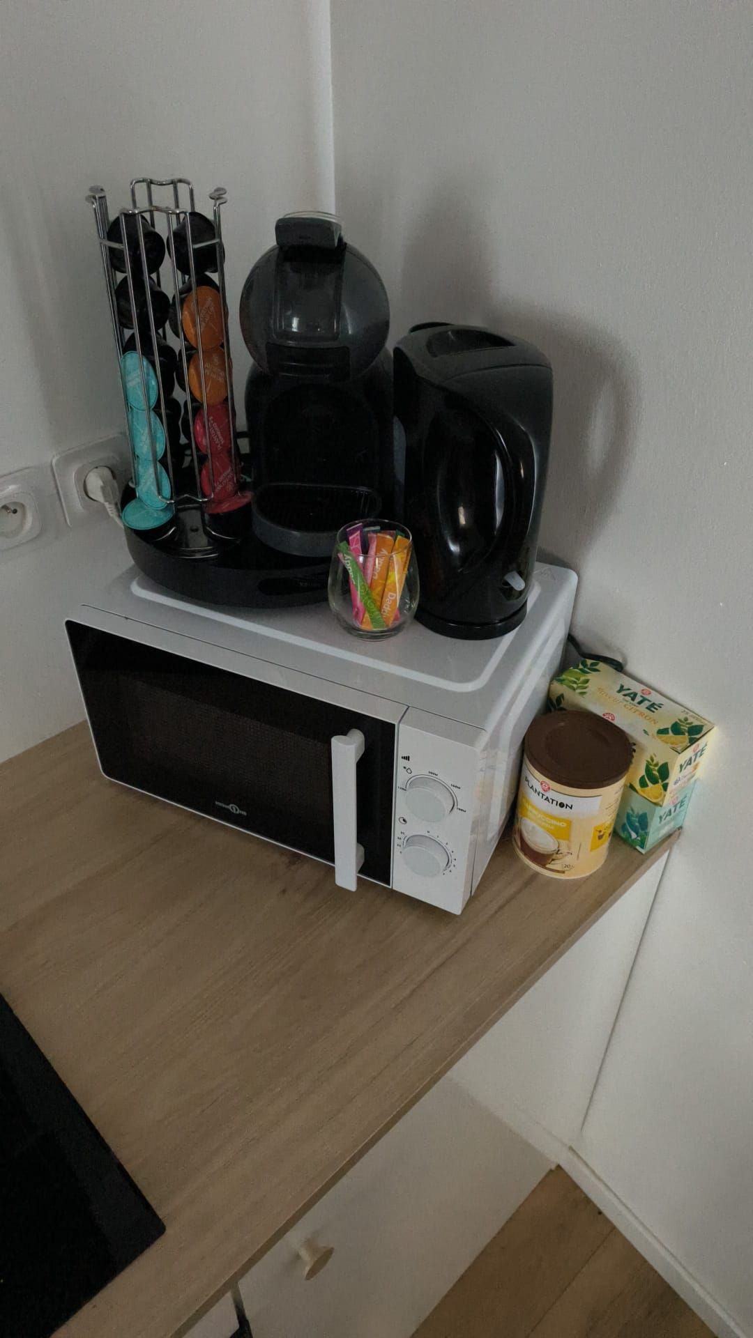 Een magnetron op een houten aanrecht, met een koffiezetapparaat, een mok, een suikerpot en een pak YATE instantkoffie.