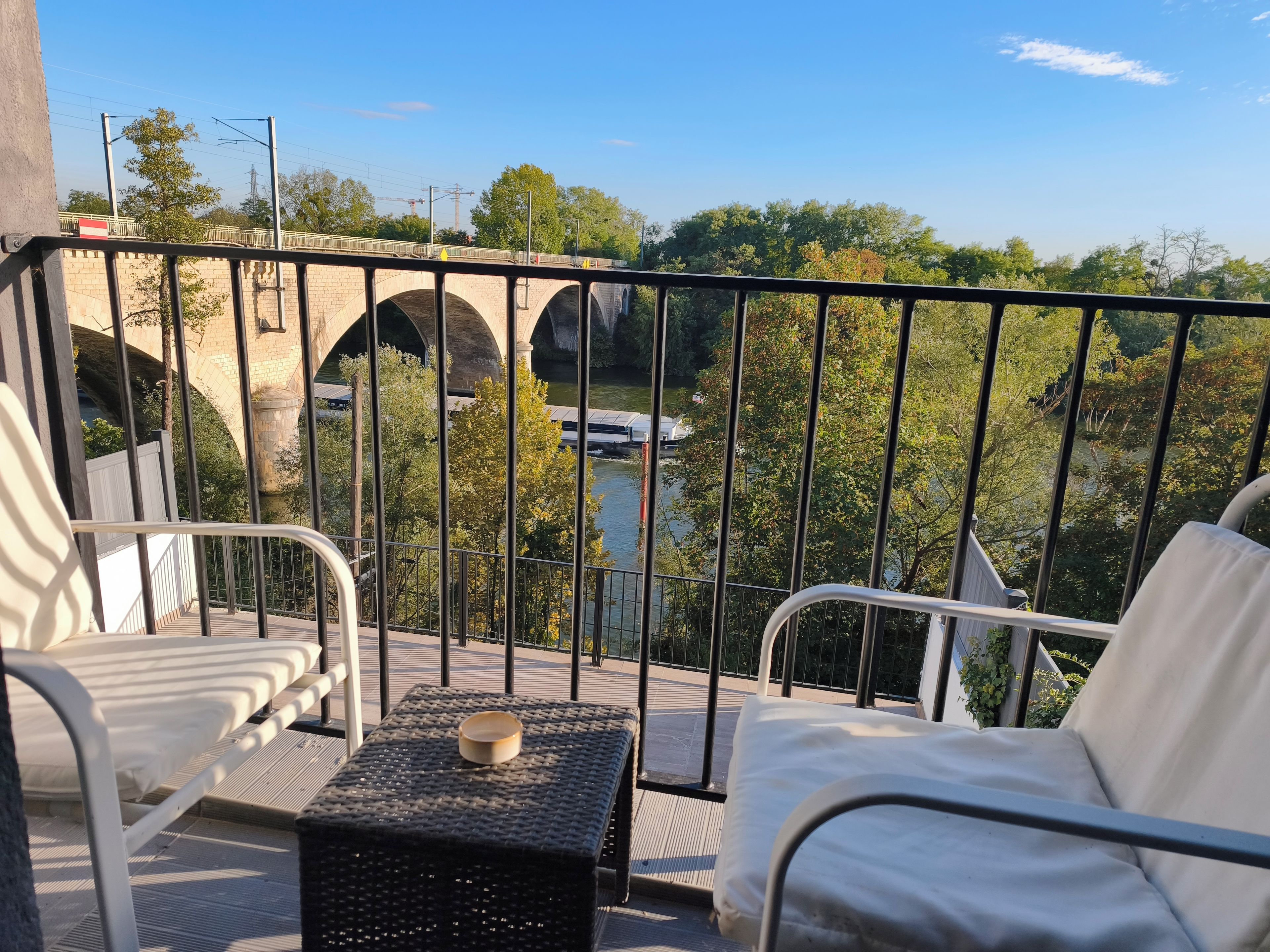 A balcony with a view of a bridge and river, furnished with two white chairs and a small table.