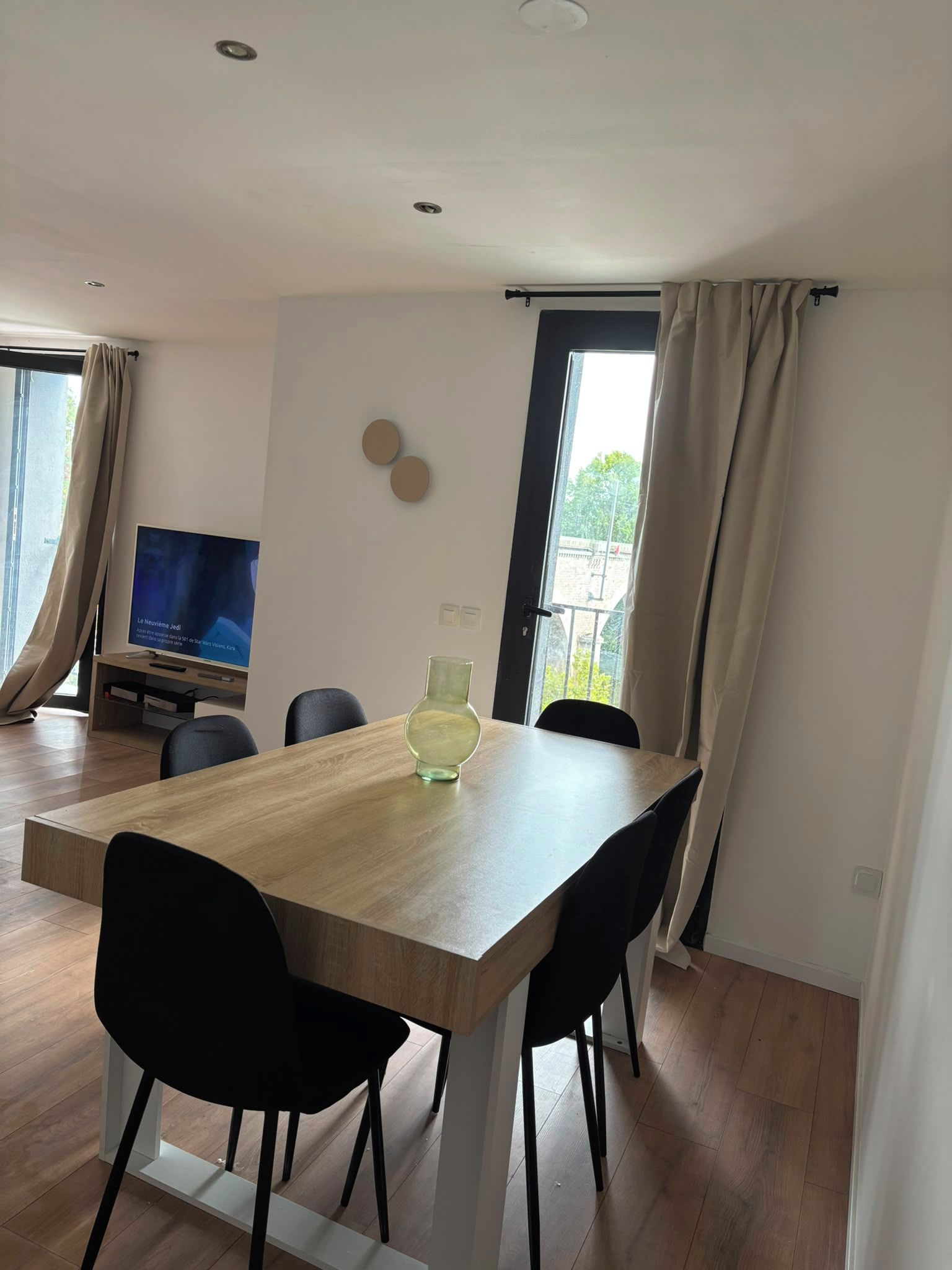Een moderne eethoek met een houten tafel, zwarte stoelen, een glazen vaas en een televisie op een standaard.