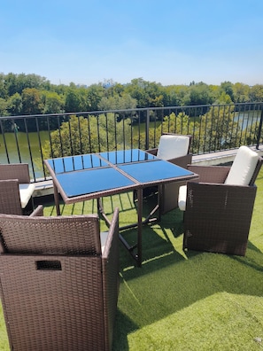Eine Terrasse im Freien mit Korbmöbeln und einem Glastisch, mit Blick auf ein Gewässer und viel Grün.
