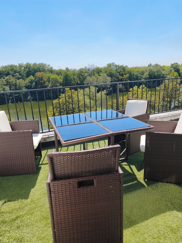 Ein Außenbereich mit Sitzgelegenheit, Tisch und Stühlen, mit Blick auf ein Gewässer und Grünflächen.