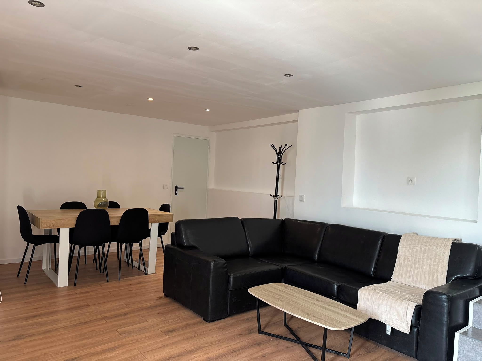 Een moderne woonkamer met een zwarte leren bank, een houten eettafel met stoelen en een kapstok.