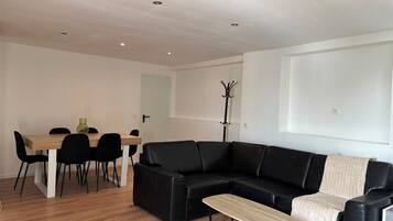 Een moderne woonkamer met een zwarte leren bank, een houten eettafel met stoelen en een kapstok.
