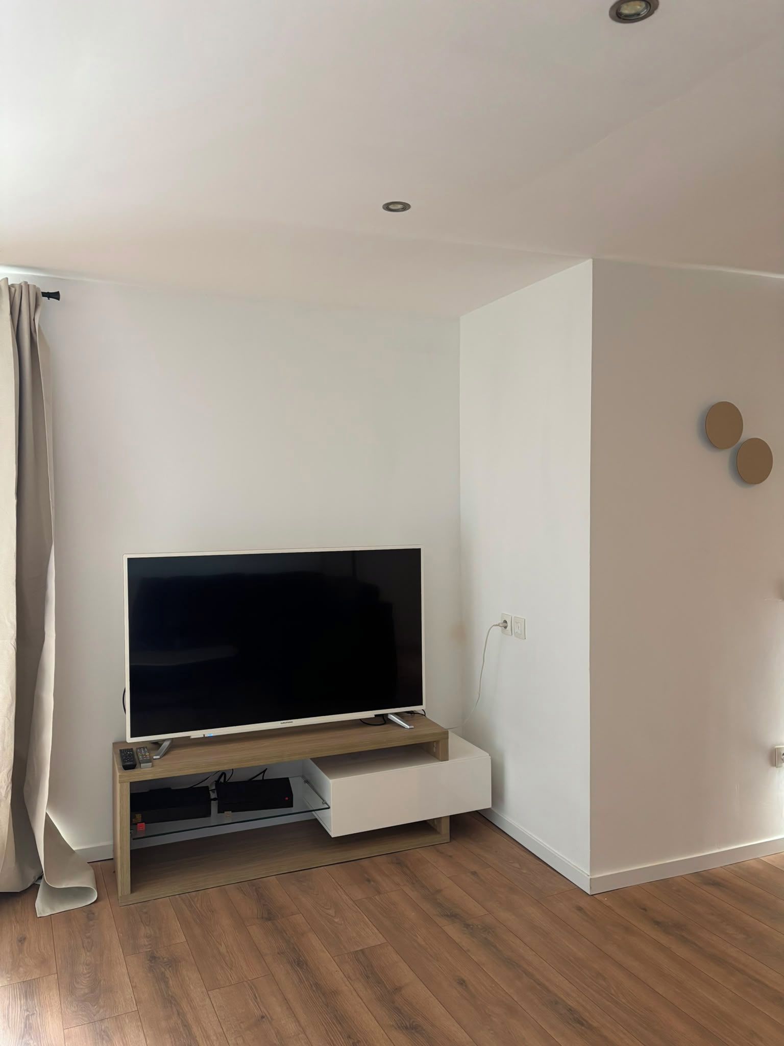 A modern living room with a flat-screen TV on a wooden stand, a white wall, and a wooden floor.