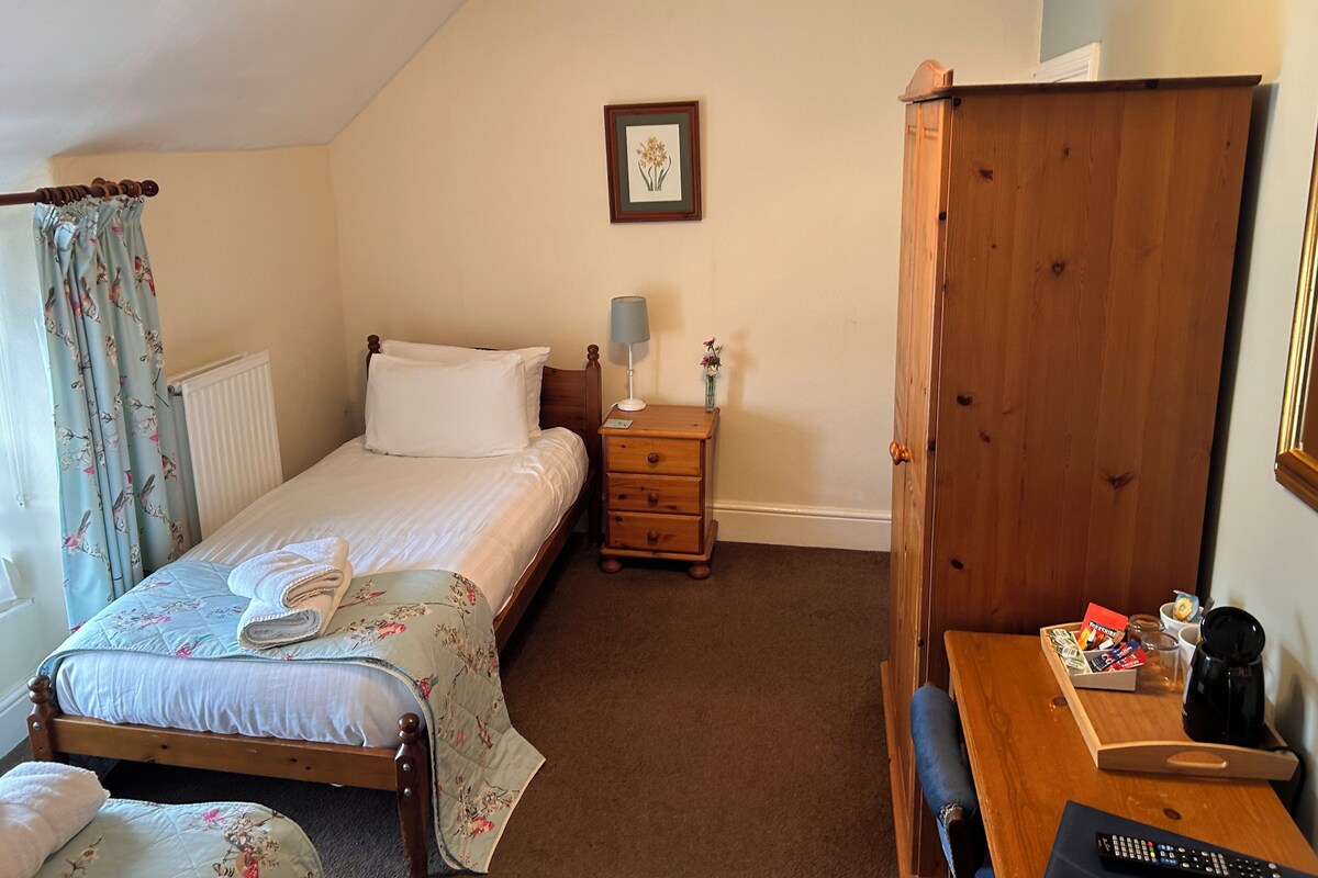 A bedroom with a bed, a bedside table with a lamp, a wooden wardrobe, and a small table with a coffee maker and remote control.