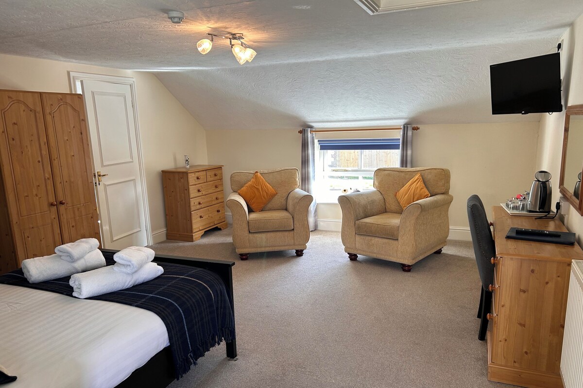 A bedroom with a bed, two armchairs, a wooden dresser, a television, and a window with curtains.