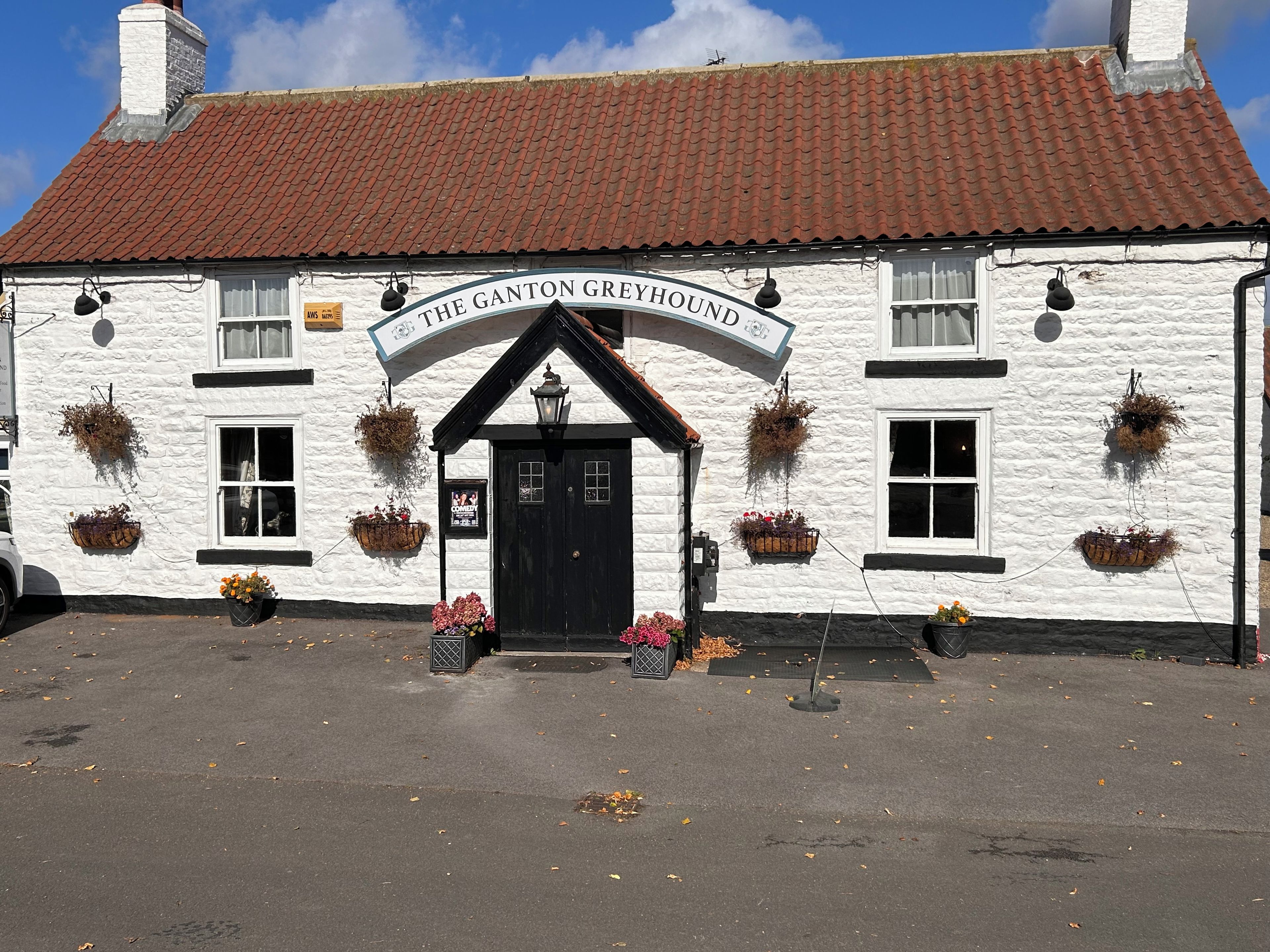 A white building with a black door and a sign reading 'The Ganton Greyhound'.