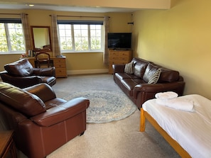 A room with a brown leather sofa, a matching armchair, a wooden bed, a TV, a dresser, and a large window with curtains. - The Ganton Greyhound (Scarborough)
