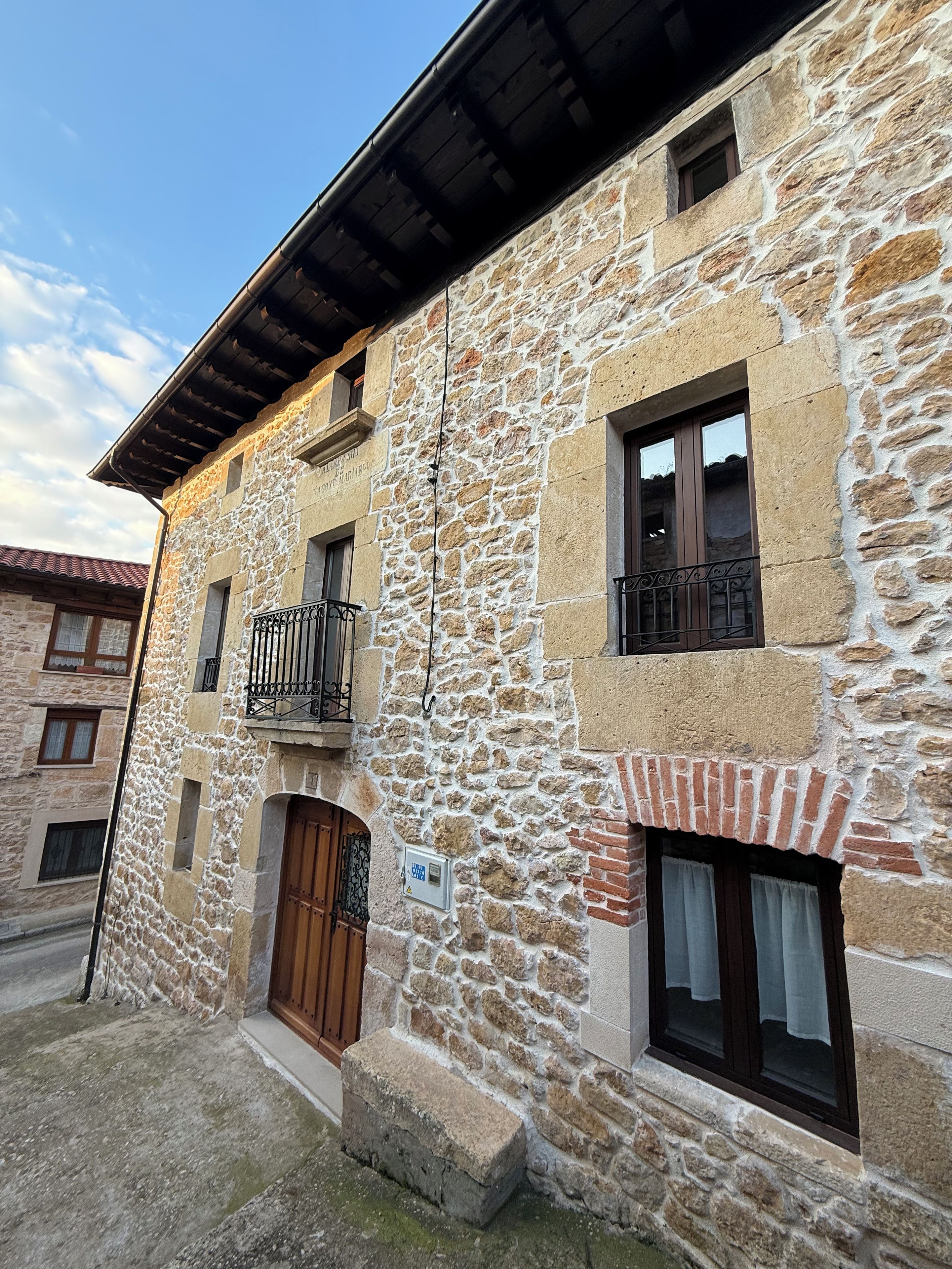 Ein Steinhaus mit einer Holztür, einem kleinen Balkon mit Metallgeländern und einem Fenster mit Vorhang.