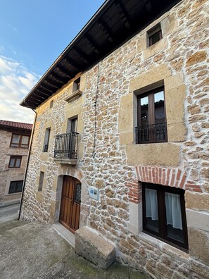 A stone building with a wooden door, a small balcony with metal railings, and a window with a curtain.