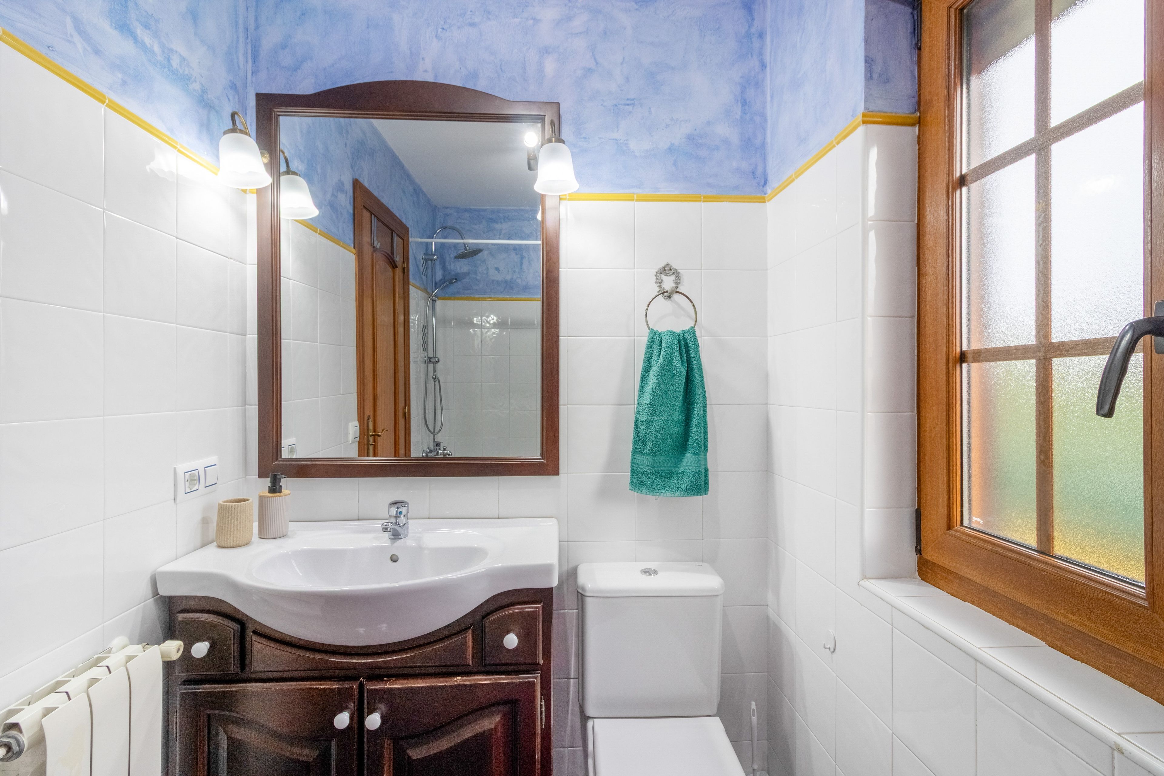 Un baño con lavamanos blanco, un espejo, inodoro y ventana.