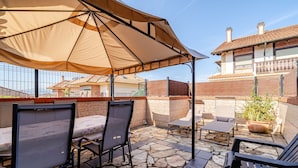Eine Terrass mit Steinfliesen, Metallstühlen, einem Tisch und einem großen Sonnenschirm, der Schatten spendet.