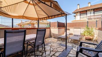 Un patio al aire libre con suelo de piedra, sillas de metal, una mesa y una sombrilla grande que proporciona sombra.