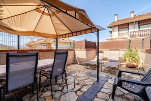 Een terras buiten met stenen vloer, metalen stoelen, een tafel en een grote parasol voor schaduw.