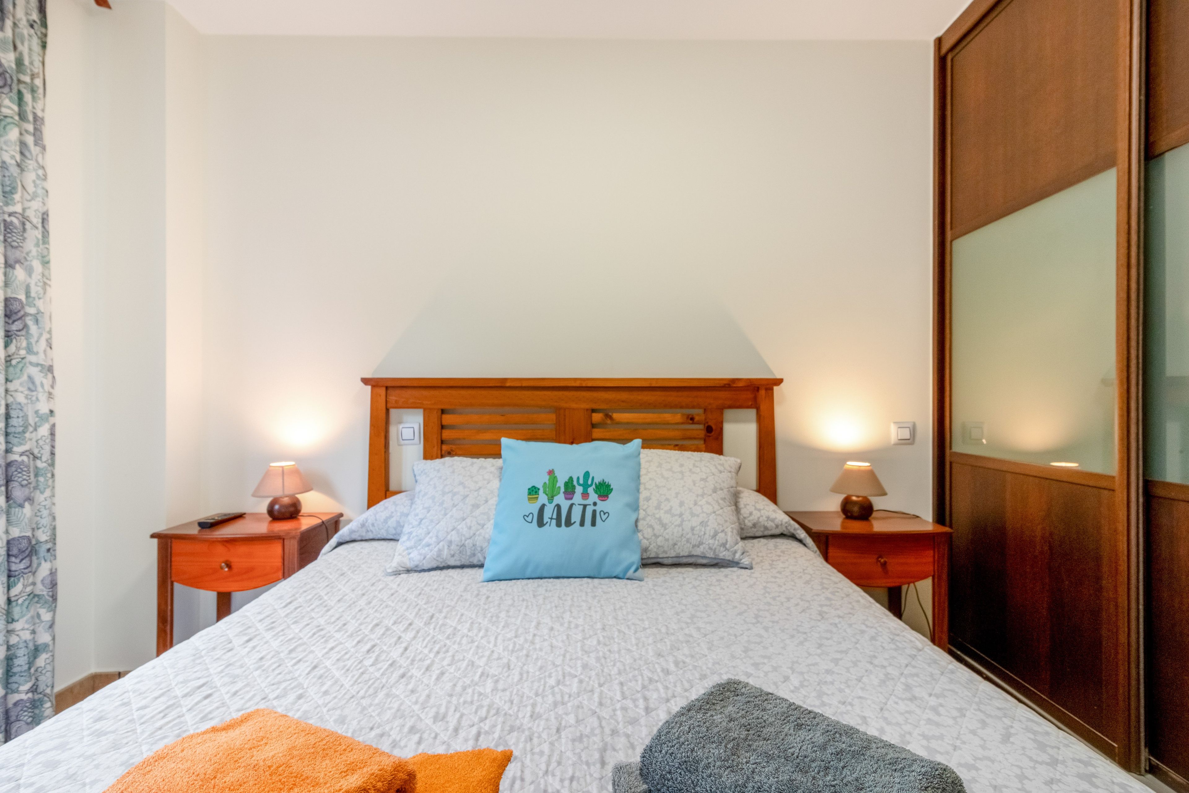 A bedroom with a wooden headboard, two bedside lamps, a bed with a quilted cover, and a decorative pillow with a cactus design.