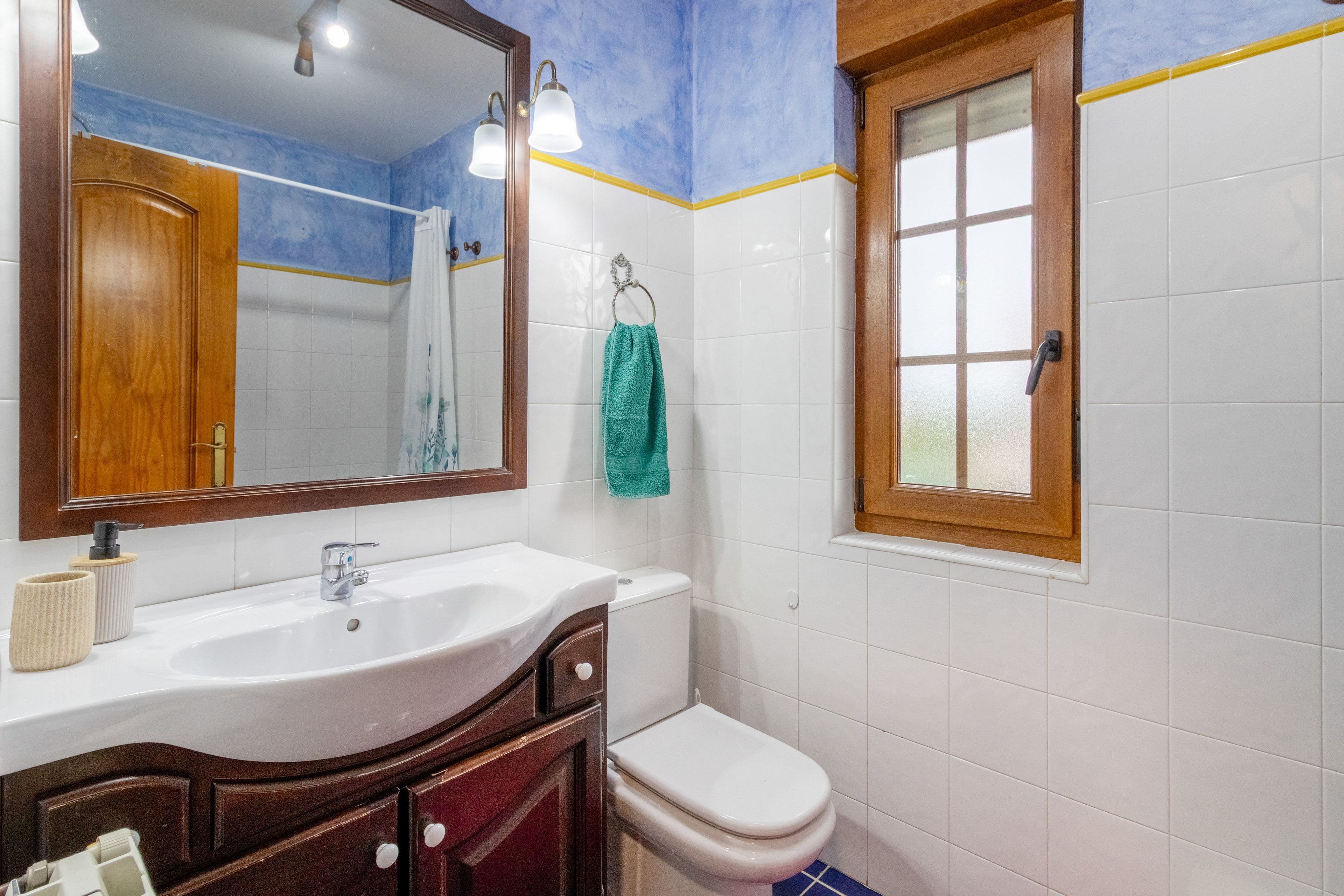 Un baño con un lavamanos blanco, tocador de madera, un inodoro, una toalla colgada en un perchero y una ventana con marco de madera.