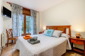 Un dormitorio ordenado con cama, sillón, mesa, televisor y ventana con cortinas.