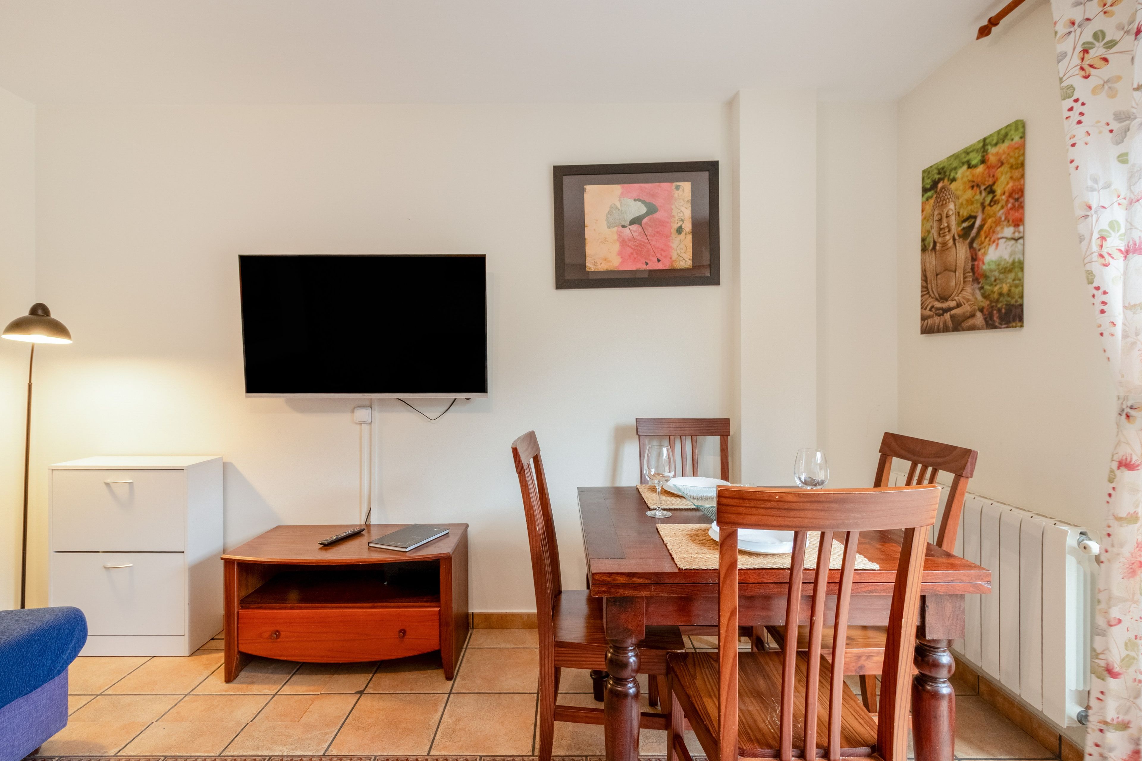 Un comedor con mesa de madera, sillas, televisor en la pared, un mueble bajo y un sofá azul.