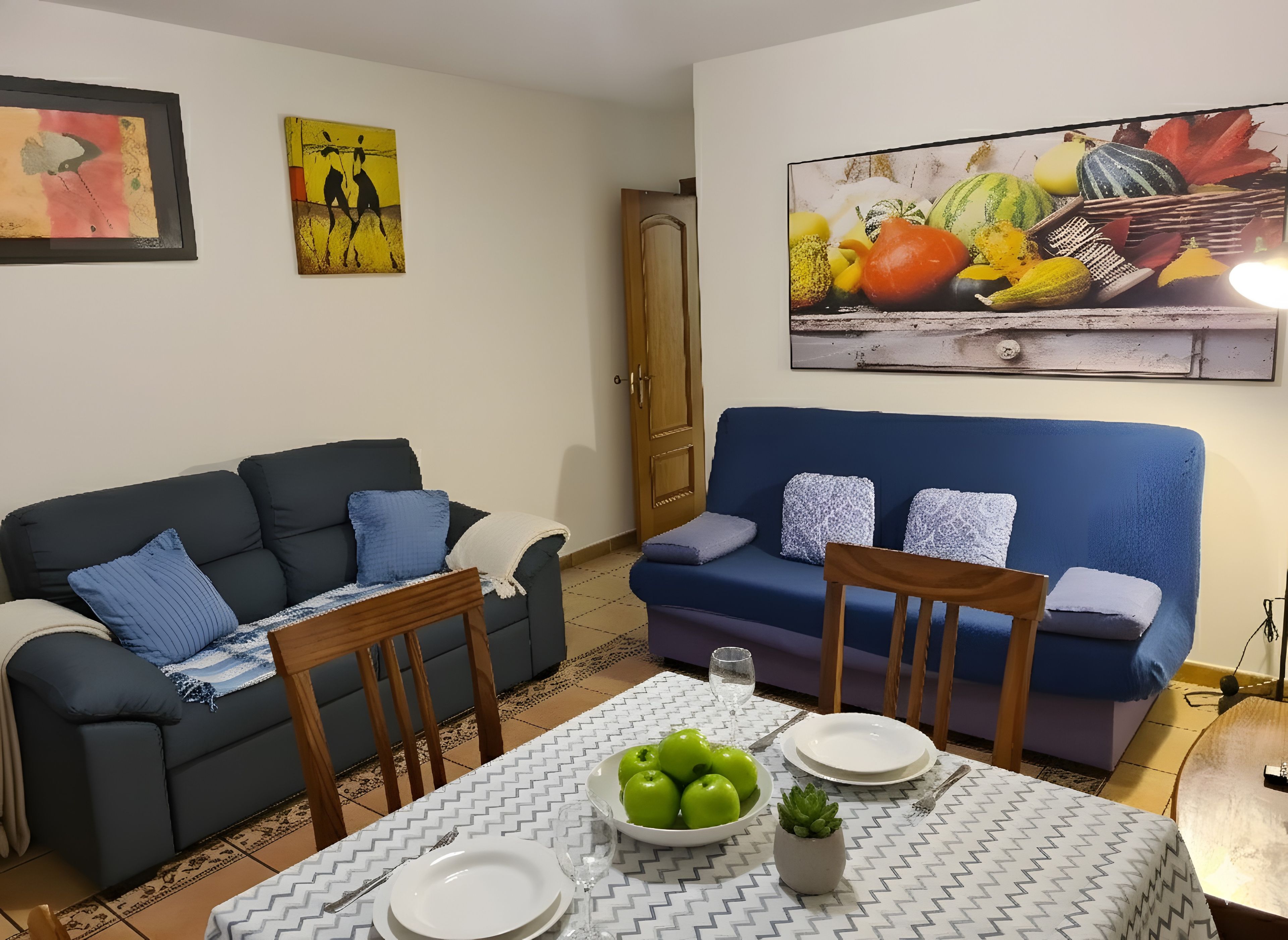 Una sala de estar con dos sofás, una mesa de comedor con platos y un tazón de manzanas, y un cuadro en la pared.