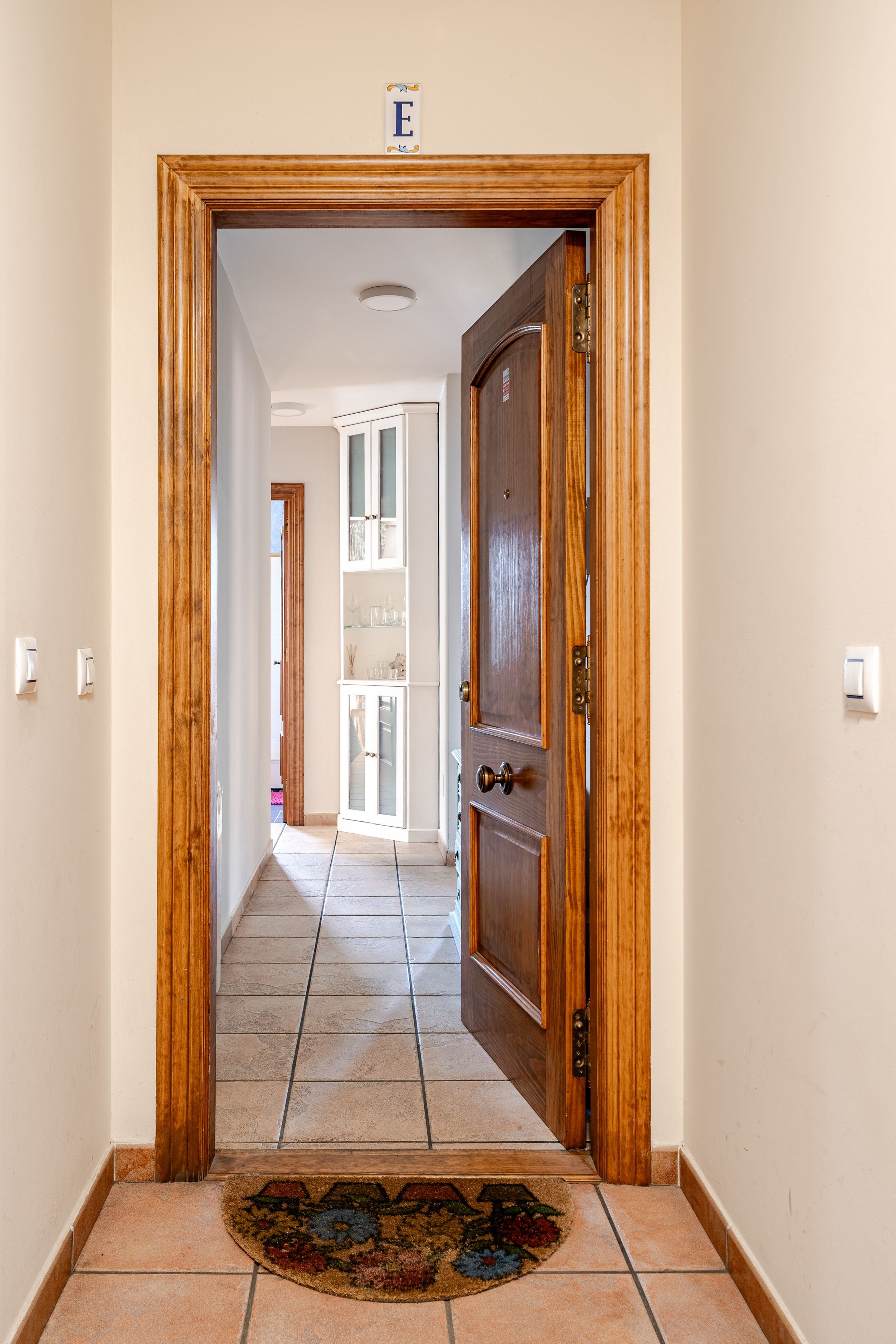 Un pasillo con puerta de madera, piso de baldosas y un tapete en la entrada.
