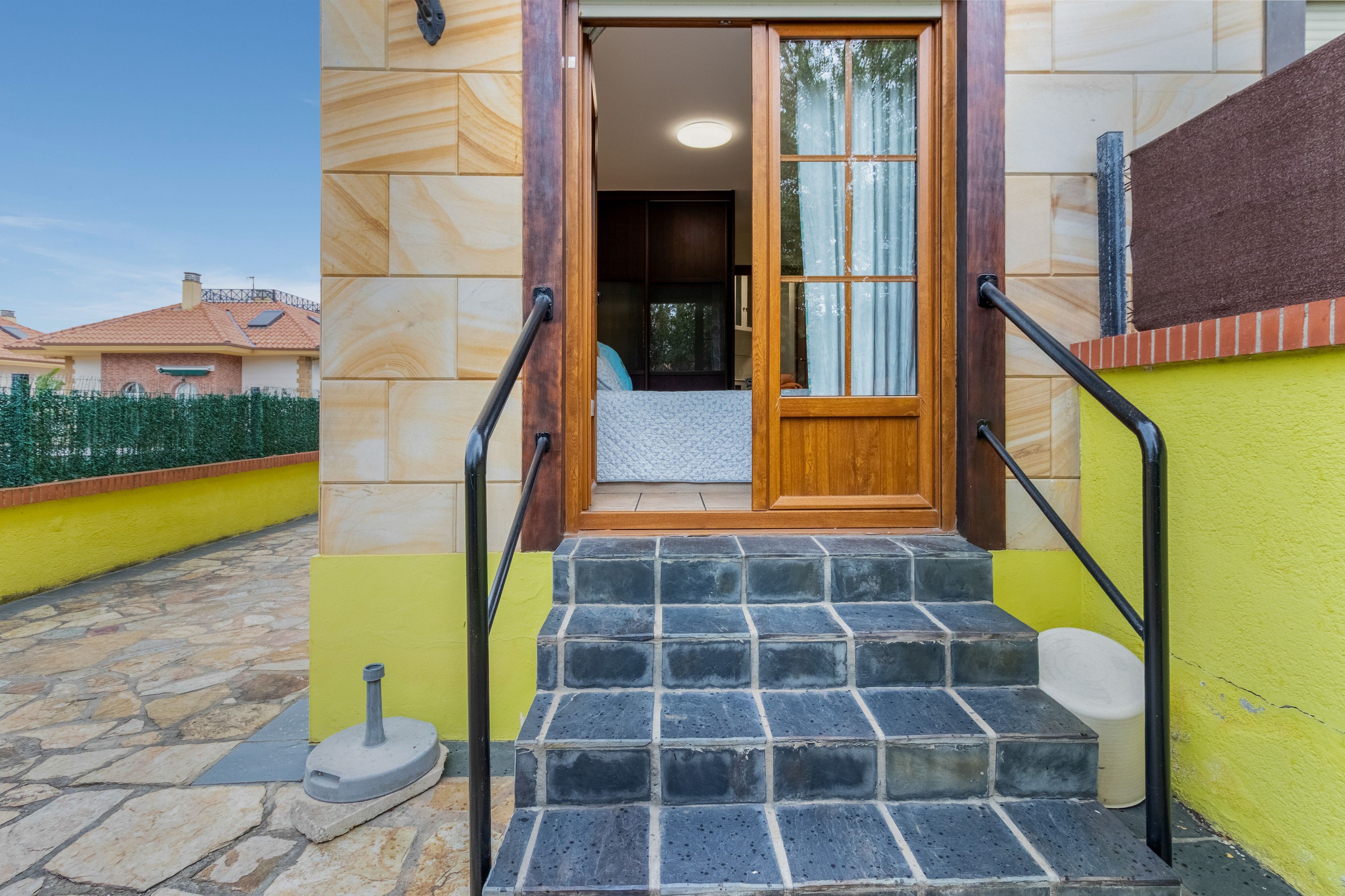 A set of stone steps leading to a wooden door with a glass panel, a metal handrail on each side, and a small outdoor area with a stone floor.