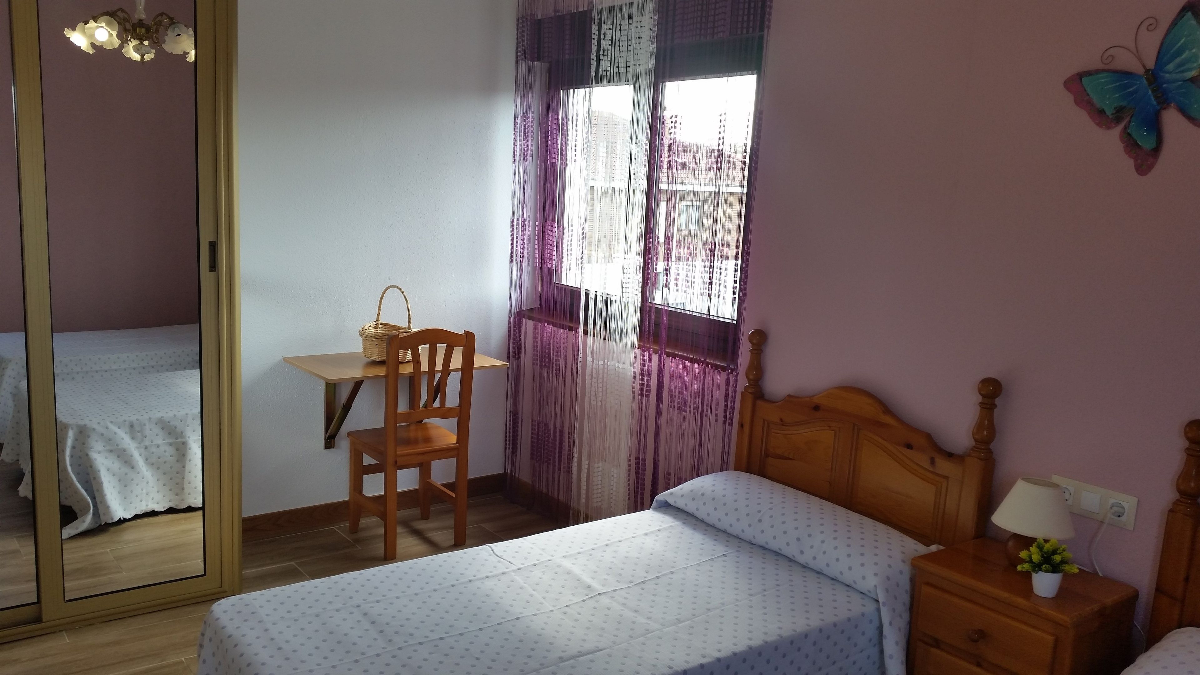 A bedroom with a bed, a wooden chair, a small table with a basket, a nightstand with a lamp, and a window with curtains.
