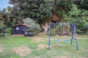 A playground with swings and a small building, surrounded by trees and a rocky cliff.