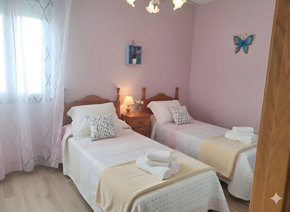 A bedroom with two beds, a nightstand with a lamp, a wall decoration, and a window with sheer curtains.