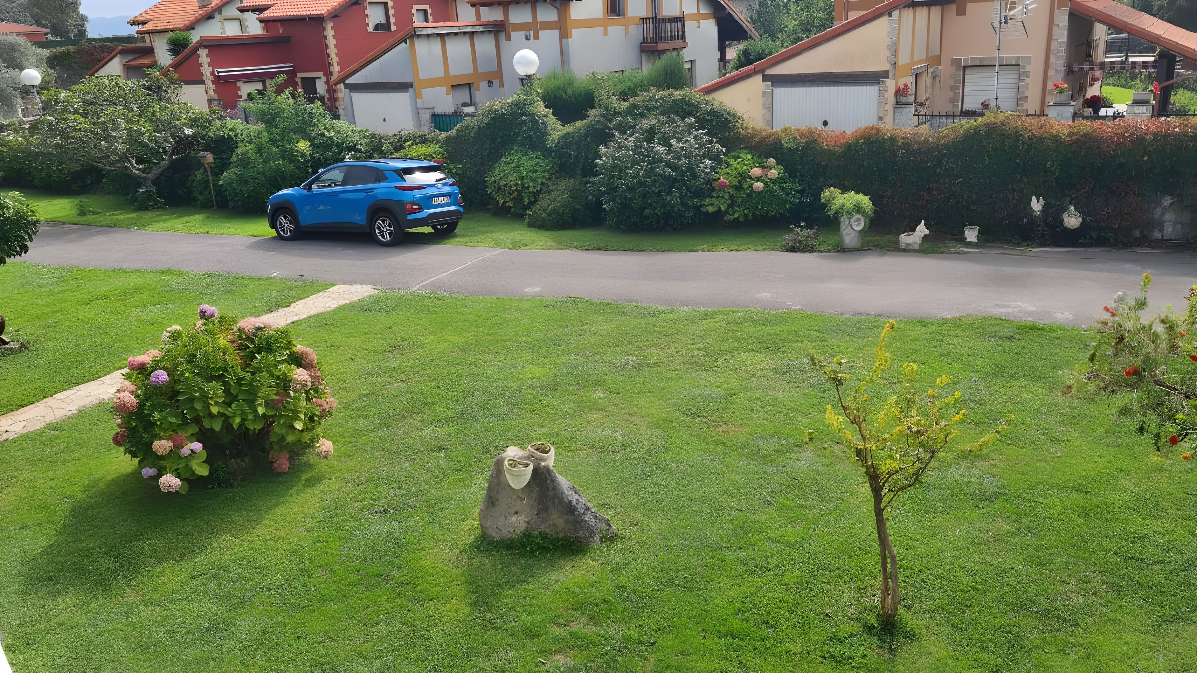 A residential area with a blue car parked on the street, well-maintained lawns, and a variety of shrubs and flowers.