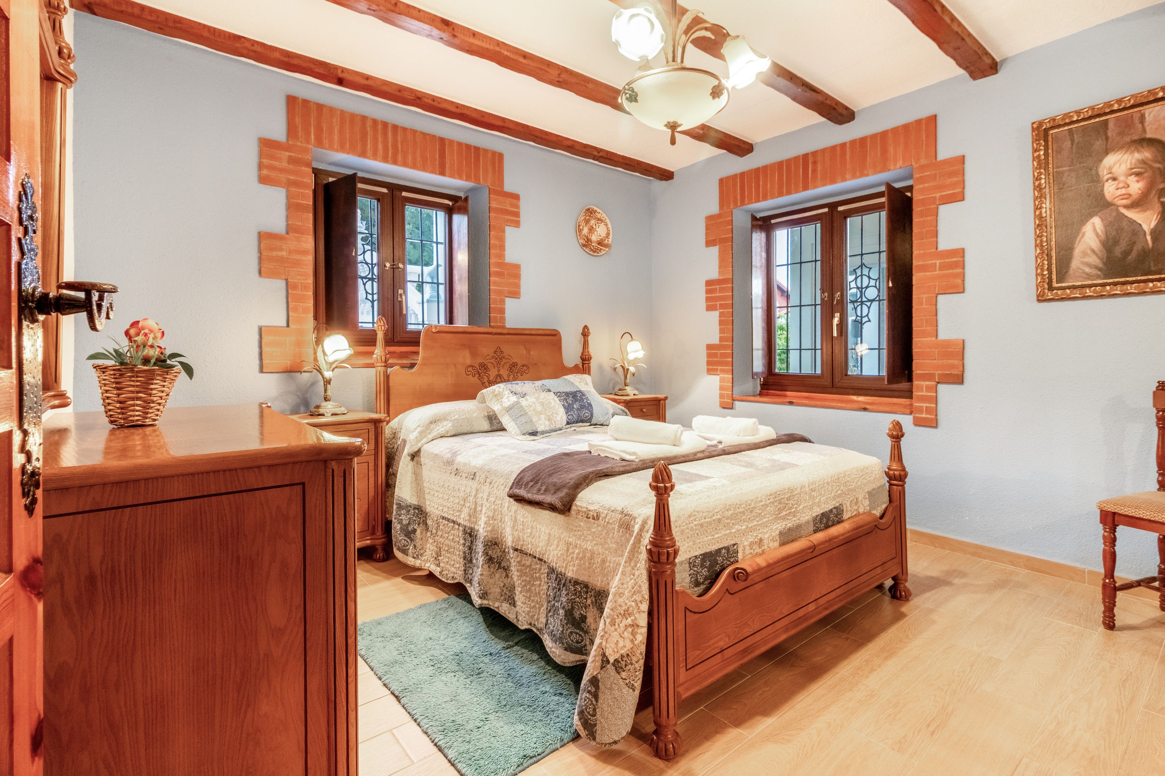 Een slaapkamer met een houten bed, een dressoir, een nachtkastje, een lamp en een lijst met een afbeelding aan de muur.