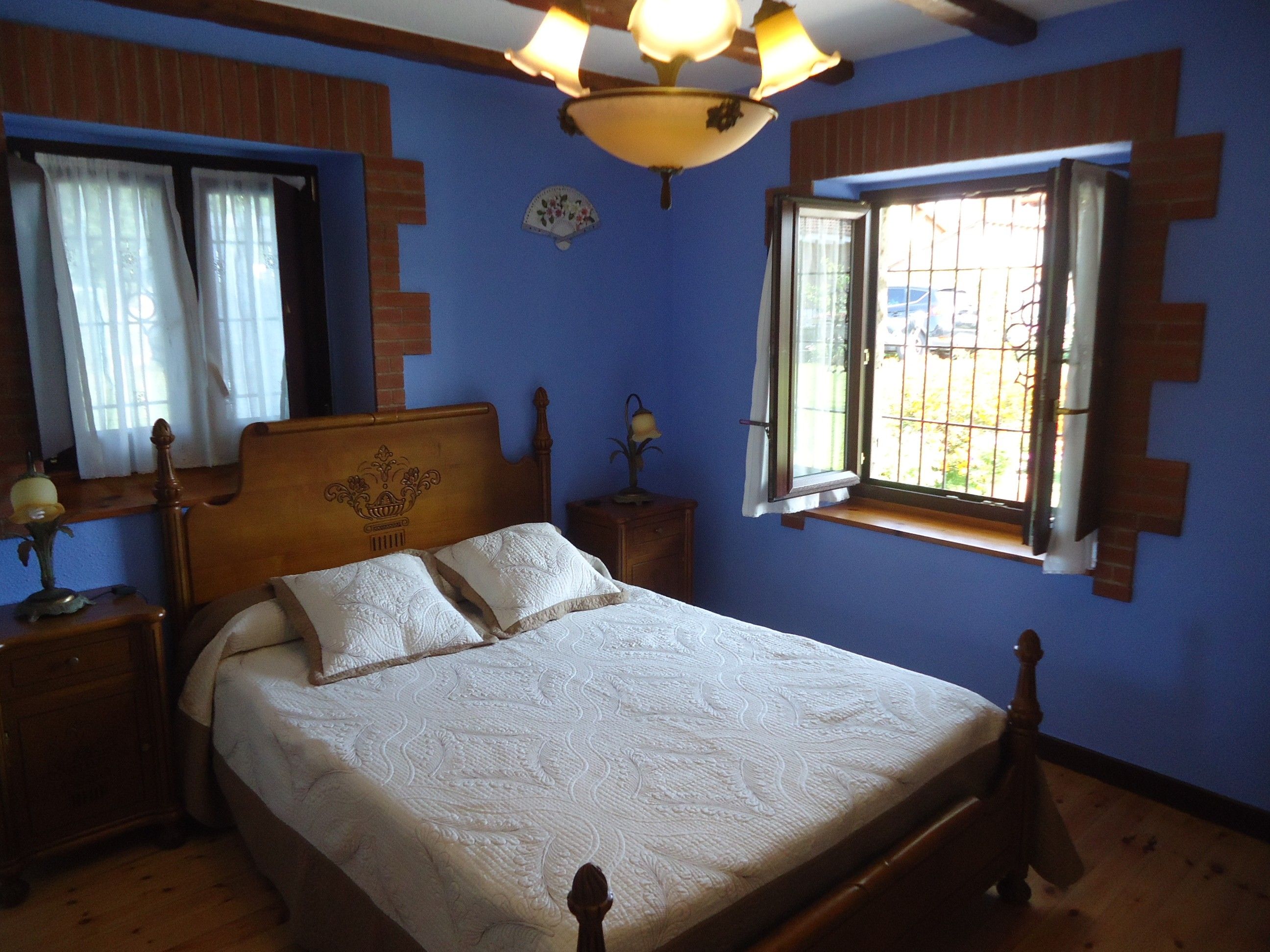 A bedroom with a wooden bed, a nightstand, a lamp, a window with bars, and a ceiling fan.