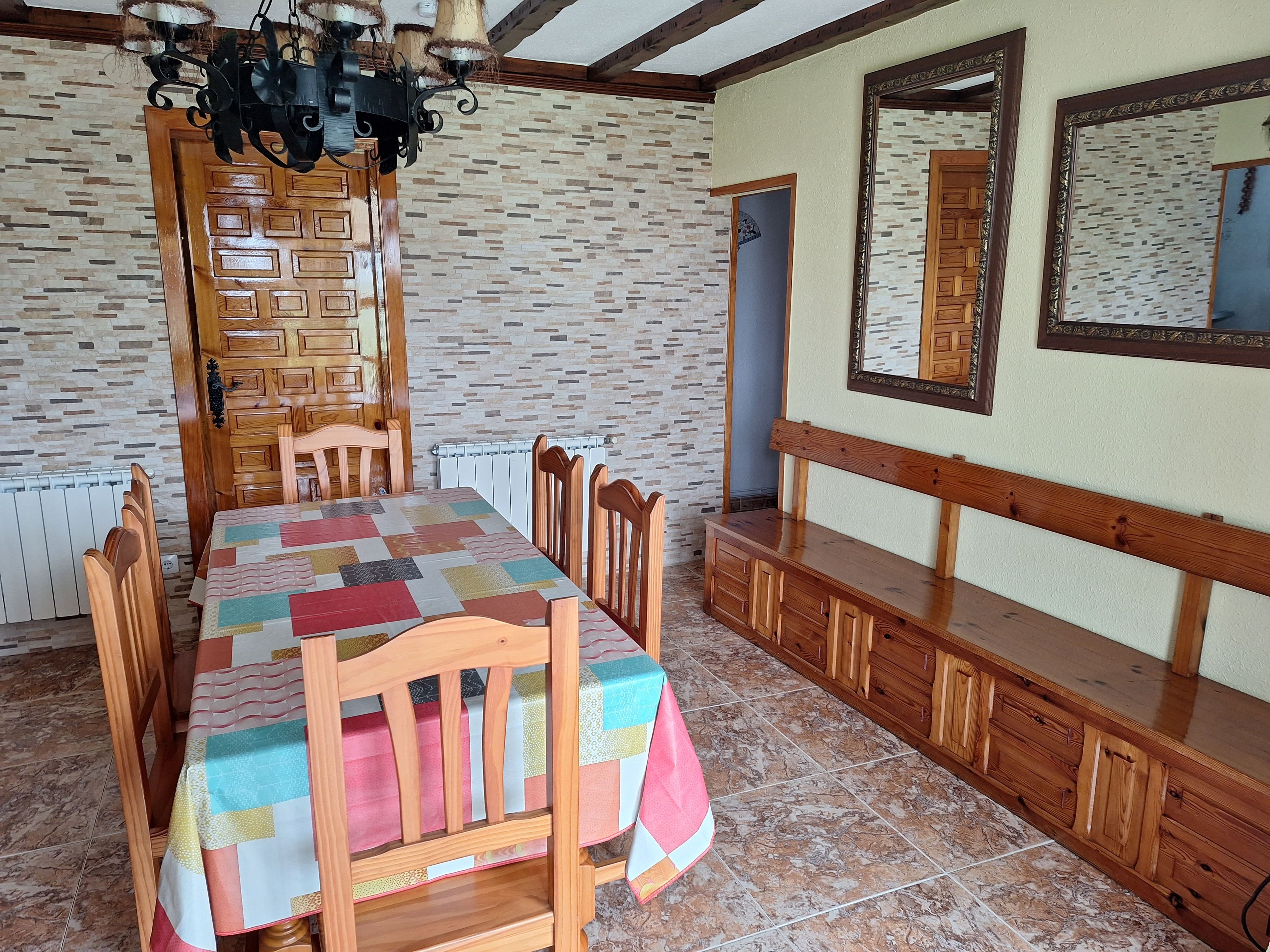 A dining area with a wooden table, chairs, a patterned tablecloth, a chandelier, and a mirror on the wall.