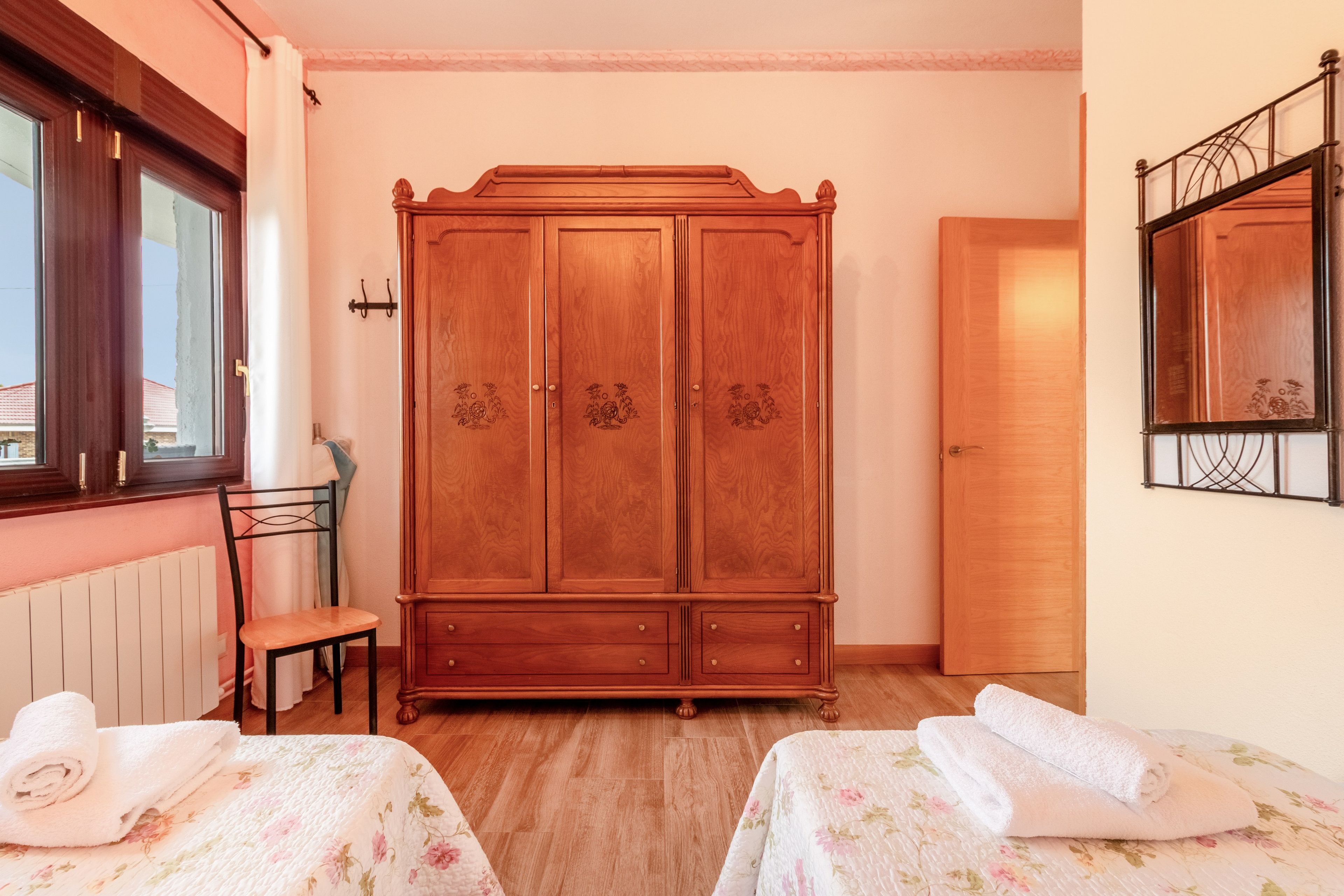 A room with a wooden wardrobe, a chair, two beds with floral bedding, a radiator, and a window with curtains.