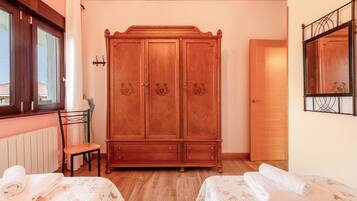 A room with a wooden wardrobe, a chair, two beds with floral bedding, a radiator, and a window with curtains.