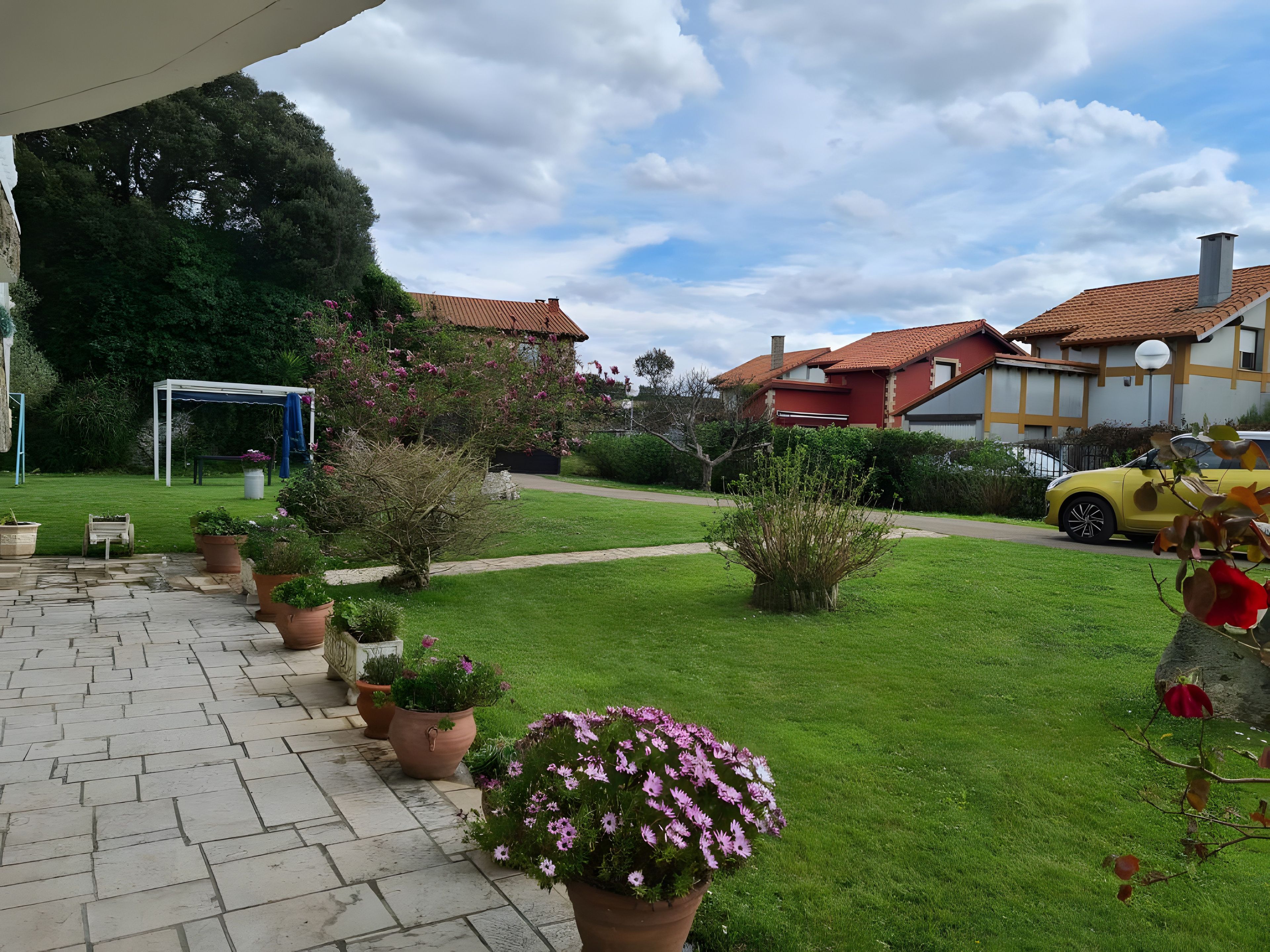 A residential area with a paved patio, potted plants, a lawn, and a yellow car parked on the street.