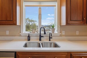 A kitchen with a double sink, wooden cabinets, a window with a view, and a potted plant on the windowsill. - White Horse Overlook | Lancaster | Amish Country (Gap)