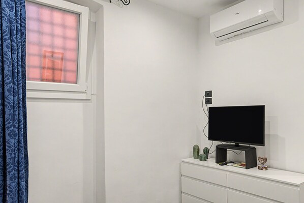 A room with a white wall, a window with blue curtains, a flat-screen TV on a white dresser, and a wall-mounted air conditioner.