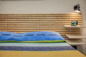 Een bed met een deken met blauw en groene patronen, een houten lattenkap, een nachtkastje met een lamp en een klein plankje met een decoratief object.