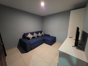Et værelse med en hjørnesofa, et fladskærms-tv og et hvidt skrivebord.