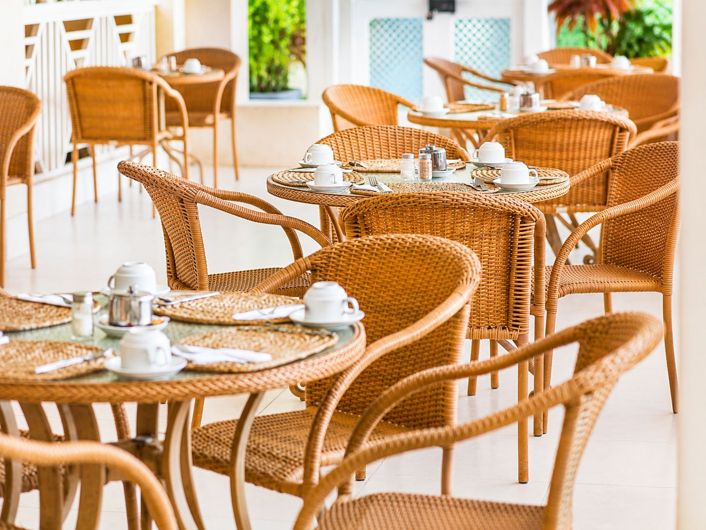 A series of wicker tables with chairs, each set with white cups and saucers, suggesting a dining or tea area.