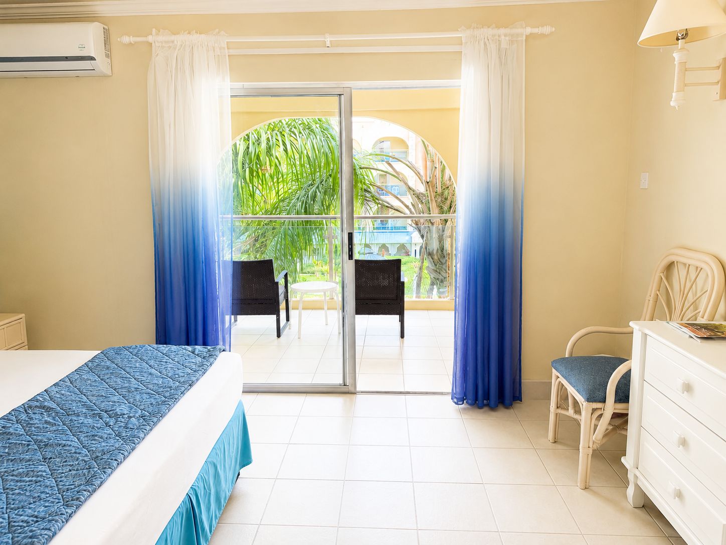 A hotel room with a bed, a dresser, a chair, and a balcony with curtains.