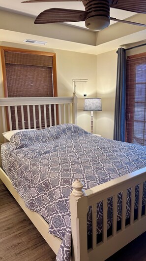 Ein Schlafzimmer mit Bett, Deckenventilator, Fenster mit Jalousien und Nachttisch mit Lampe.