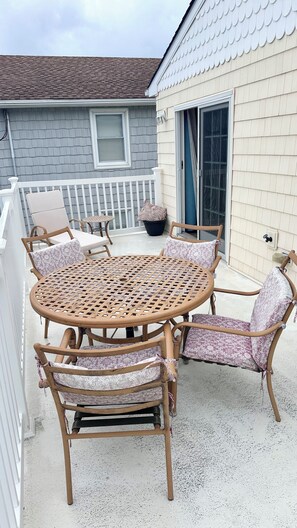Eine überdachte Terrasse im Freien mit einem runden Tisch und Stühlen, einem weißen Geländer und einem Haus mit einem grauen Schindeldach.