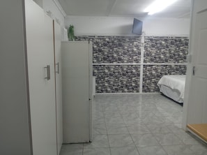 Habitació senzilla amb llit, armari blanc, nevera petita i paret amb motius de pedra.