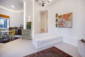 A modern living room with a white sofa, dining table with chairs, and a kitchen area in the background. - Borgo Principessa - Pool & Garden by the Sea (Bari)