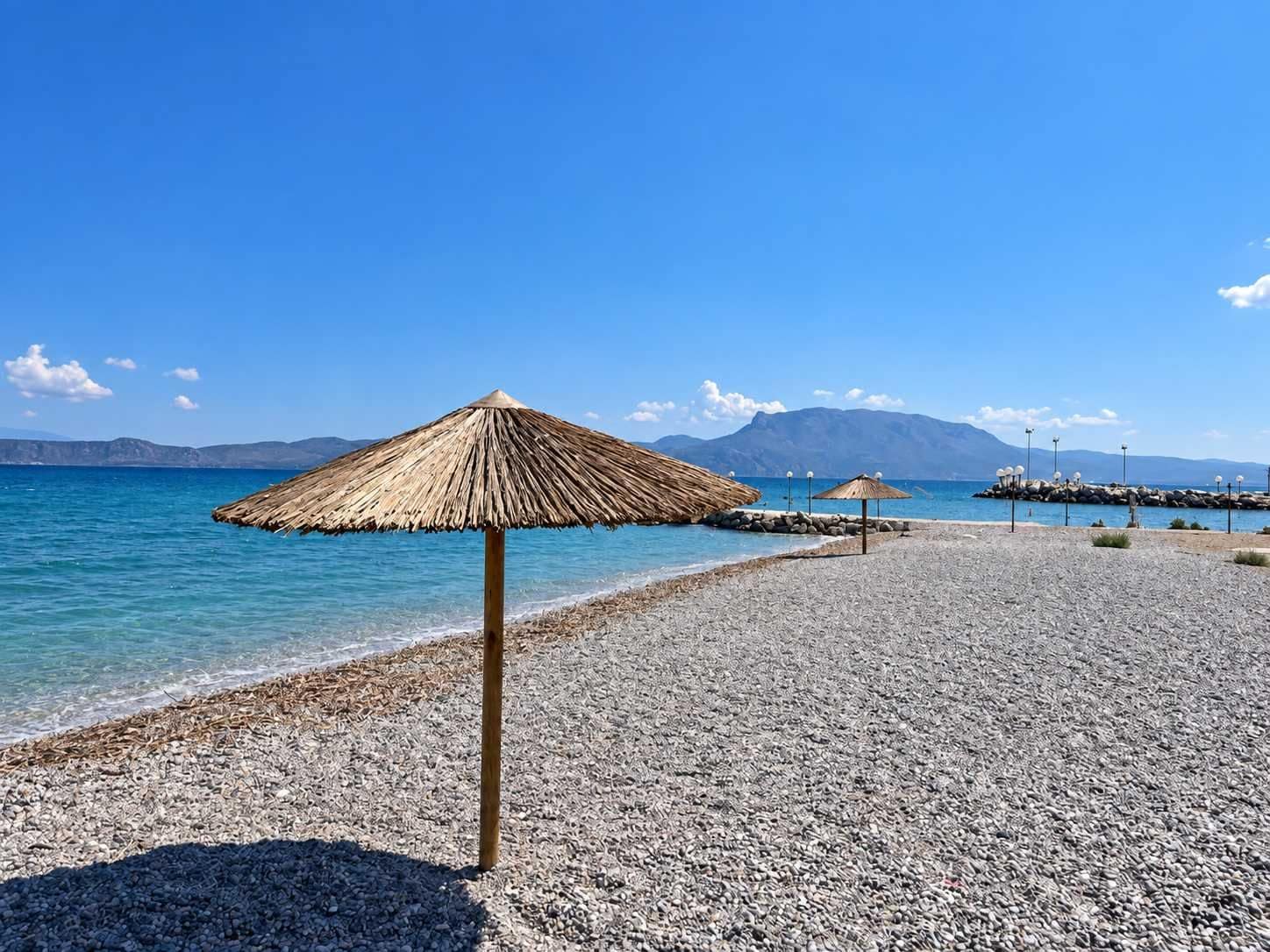 A pebble beach with a thatched umbrella and a rocky breakwater in the background.