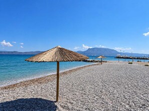 Ein Kieselstrand mit einem Strohdachschirm und einem Felssteg im Hintergrund.